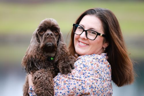 Stacie is smiling at the camera. She has a small brown cocker spaniel in her arms.